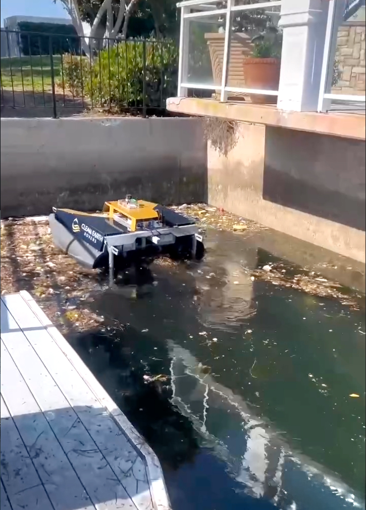 A small, autonomous surface vessel labeled "Clean Earth Rovers" is actively collecting trash and debris from a polluted waterway.