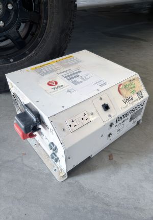 A white Volta Power Systems inverter unit with warning labels, an AC outlet, and a red battery terminal, sitting on a garage floor beside a large truck tire.