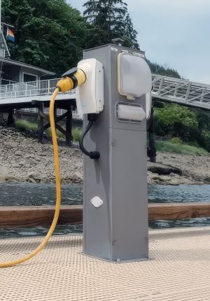 A grey marine power pedestal on a dock with a white VoltSafe magnetic connector attached to a yellow shore power cable, with a ramp and forested shoreline behind.