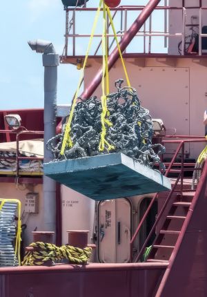 A crane on a red vessel lifting a concrete reef module with a complex latticed surface, dripping seawater, with yellow lifting straps.