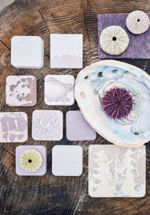 Overhead view of twelve square material samples in white, lavender, and mauve tones arranged on a weathered wood stump alongside sea urchin shells and an abalone shell holding a purple urchin.