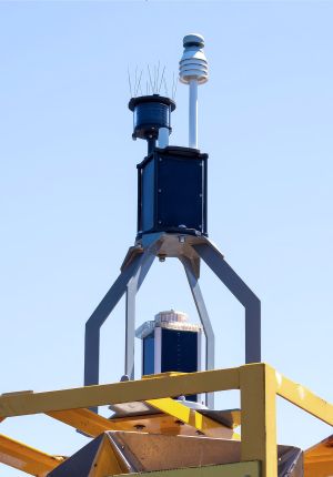 Close-up of a sensor mast on a yellow buoy frame against clear blue sky, with a weather station, camera housing, and antenna array mounted on a metal support.