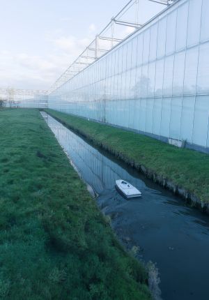 A small white autonomous surface vessel navigating a narrow drainage canal alongside the white wall of a large commercial greenhouse.