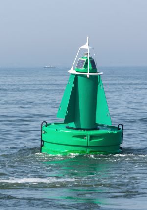 A green conical navigation buoy with solar panels and a light beacon floating on calm grey-blue sea, with a small vessel in the distance.