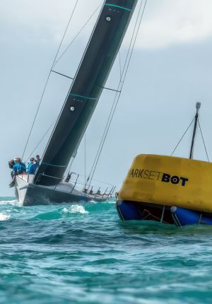 A racing sailboat heeling over with crew hiked out on the rail, passing a yellow autonomous MarkSetBot race mark buoy in choppy turquoise water.