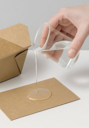 A hand tilting a glass beaker to pour a clear viscous bioplastic coating onto a sheet of kraft paper.