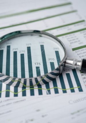 A magnifying glass resting on printed financial reports, enlarging a dark green bar chart with numeric labels and a trend line.