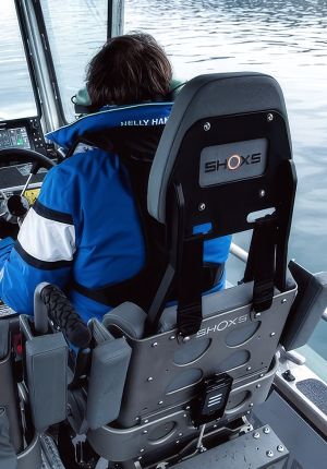 A boat operator in a blue jacket at the helm, seated beside an empty grey SHOXS shock-mitigating suspension seat on a vessel deck.