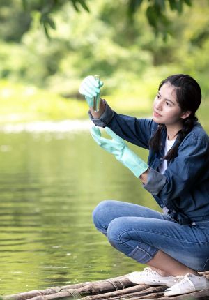 A young woman in a denim jacket and mint green gloves crouching on a wooden raft, holding up a test tube of water sampled from a green river.