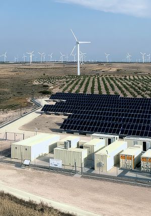 A utility-scale energy site with rows of white battery containers in the foreground, a large solar array behind, and dozens of wind turbines across the arid landscape.