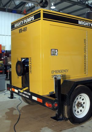A yellow trailer-mounted Mighty Pumps diesel pump unit parked inside a workshop, with labeled intake, emergency shut-off, and battery disconnect panels.