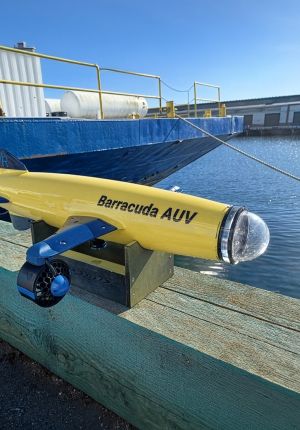 Yellow and blue 'Barracuda AUV' autonomous underwater vehicle resting on a wooden stand at a dock, with a blue support barge and clear water in the background.
