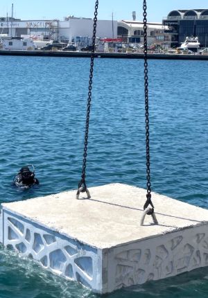 A textured concrete reef block suspended by chains being lowered into harbor water, with a scuba diver waiting nearby and a waterfront marina behind.